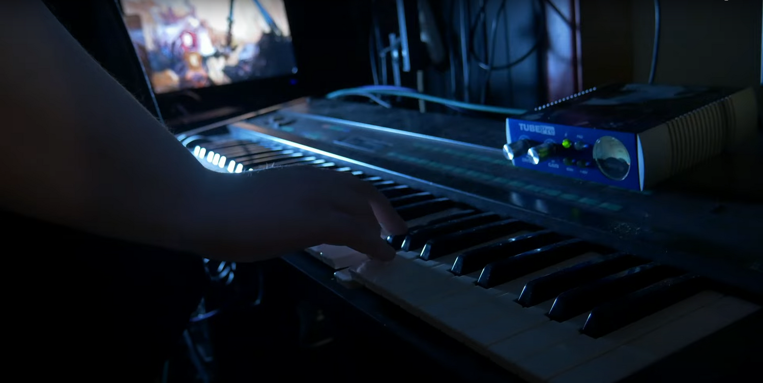 Person playing a keyboard in a dimly lit room with a laptop displaying music software.